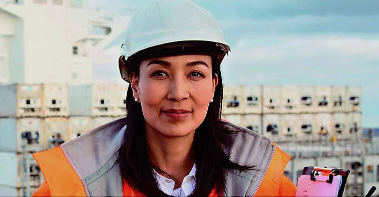 Woman wearing a hardhat with a clipboard and walkie talkie on a shipyard in front of a container ship